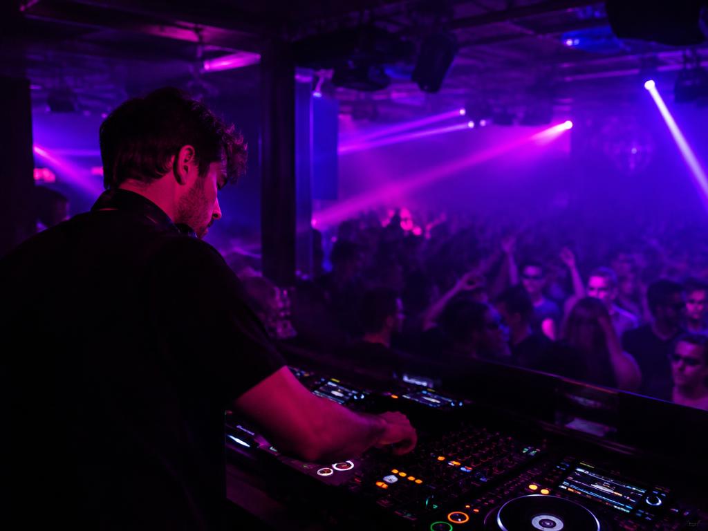 DJ mixing at the decks under magenta and violet club lights, crowd dancing behind